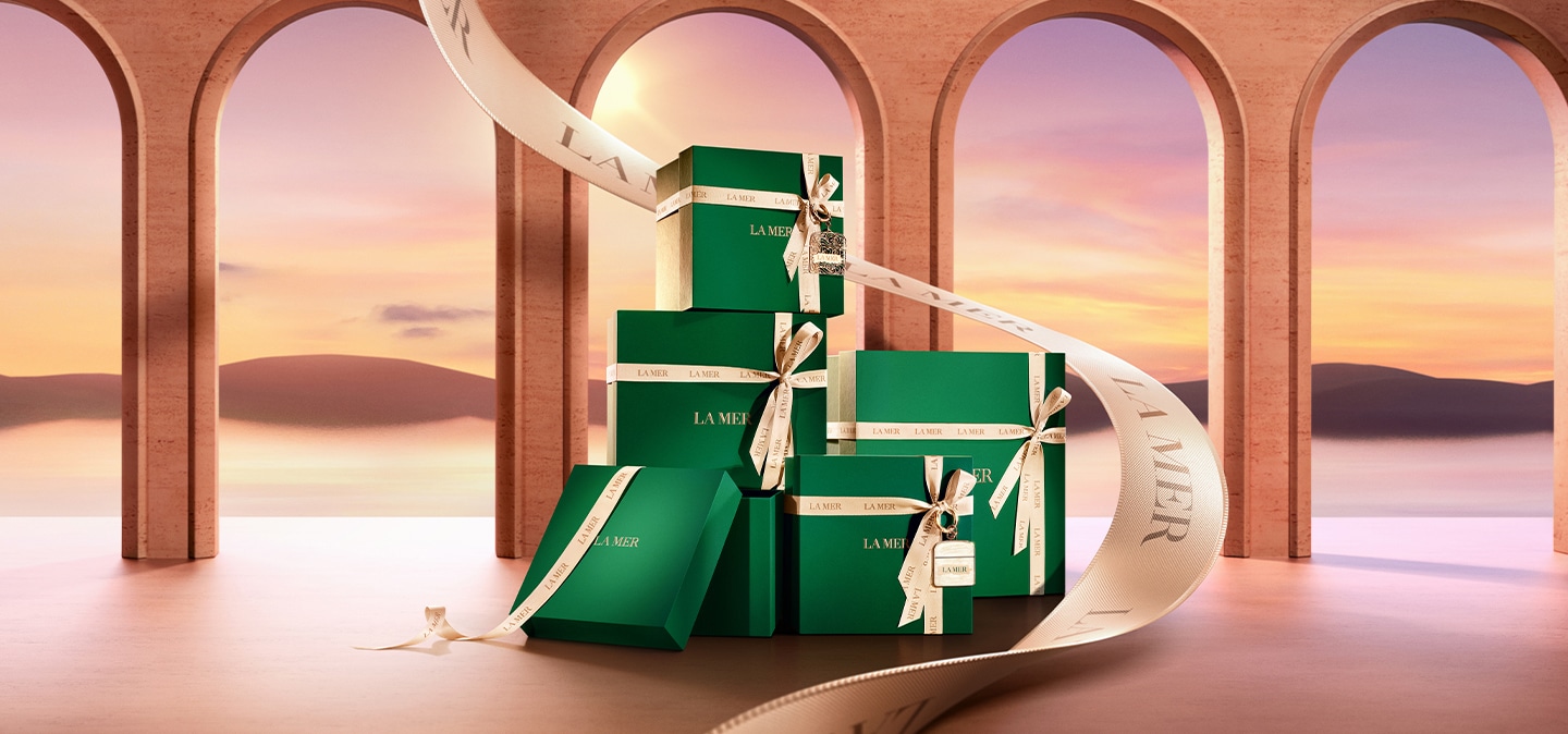 A stack of la mer gift boxes with a larger ribbon running up behind them against a background of arch windows and golden sunset.